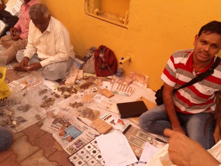 Two men sit on a sidewalk with sheets of old coins around them in Panjim, Goa.